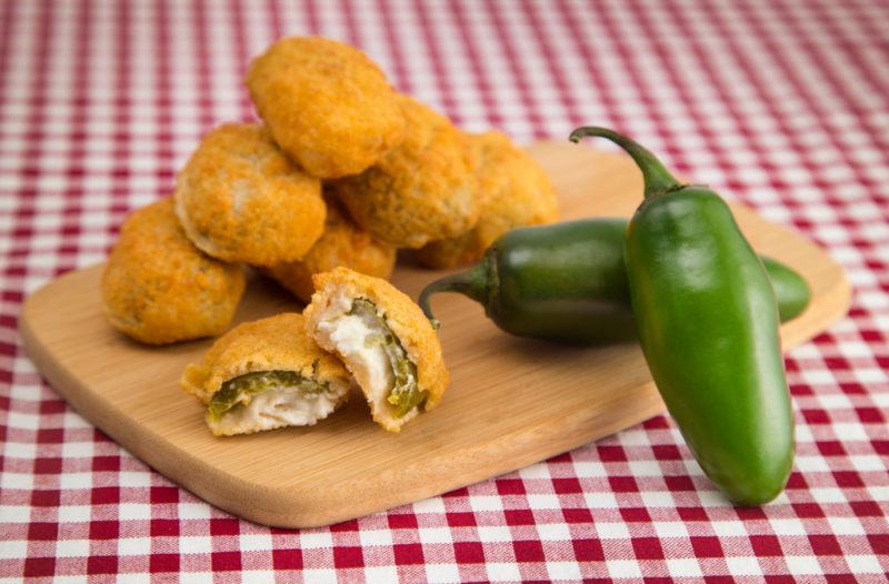 A wooden board on a red and white checkered cloth that has deep fried jalapeno poppers and two fresh green jalapenos