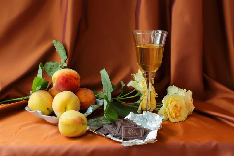 A glass of white dessert wine, some peaches, peach-colored roses, and milk chocolate