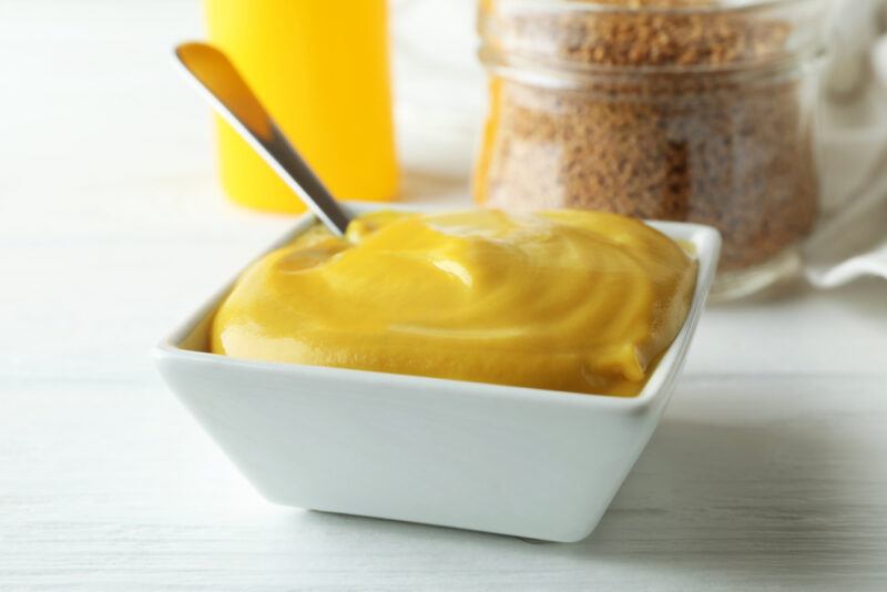 on a white surface is a square white bowl of Dijon mustard with a glass container full of mustard seeds and a yellow plastic container