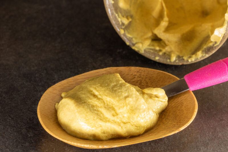 overhead shot of a wooden oval-shaped container with a spoon with pink handle full of Dijon mustard, beside it is a jar of Dijon mustard