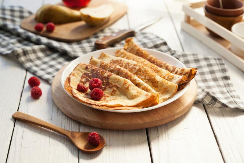 A white plate on a wooden board that has various thin pancakes and berries. The table also includes other breakfasti items, like a spoon, a cloth and more pancakes