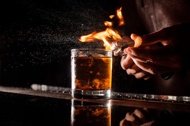 A Don Lockwood whiskey cocktail on a bar with a bartender making a flaming orange peel