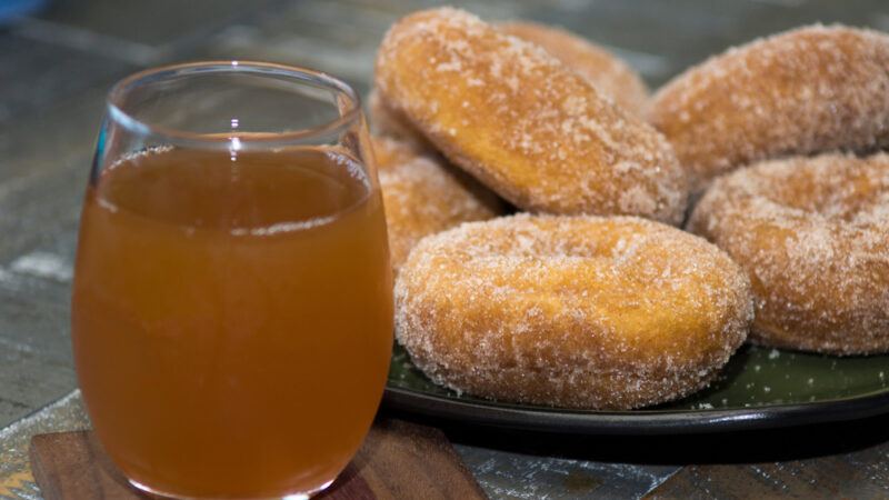 A dark plate of sugar-coated doughnuts rests on a dark tile surface near a clear glass of hot apple cider.