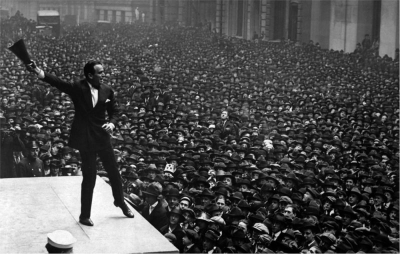 Douglas Fairbanks speaking to a large crowd while holding something in his hand, in a black and white image