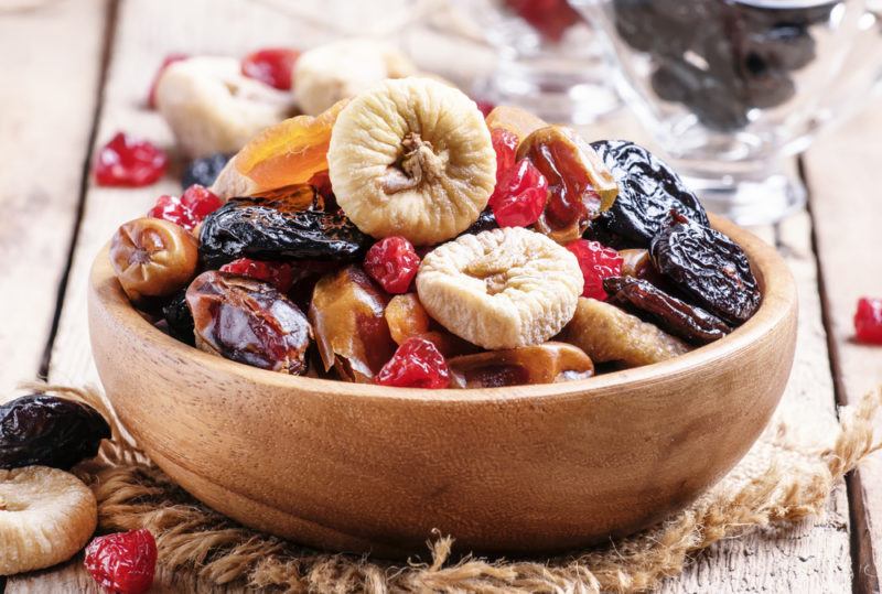 A wooden bowl on a wooden table that contains various types of dried fruit.