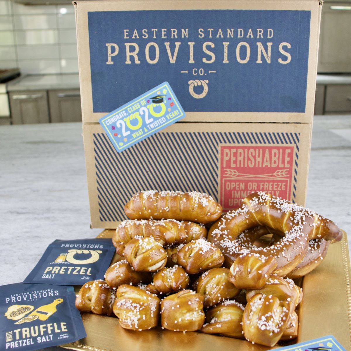 AN EASTERN STANDARD PROVISIONS CO BOX WITH A 2020 GRADUATION STICKER AND A GOLD PLATTER WITH A VARIETY OF SOFT PRETZELS AND TWO PACKS OF PRETZEL SALTS ONE PLAIN AND THE OTHER WHITE TRUFFLE.