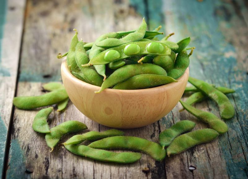 A wooden bowl of edamame on the table with edamame scattered around