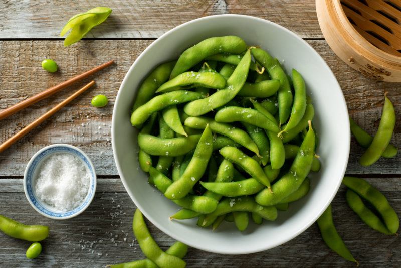 A bowl of steamed edamame with chopsticks on a wooden table