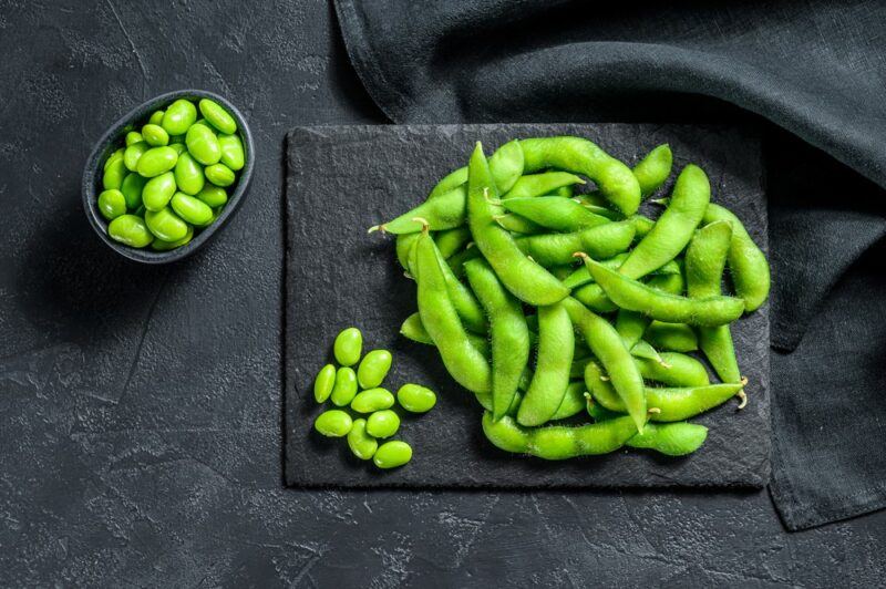 A black slate with cooked edamame beans, next to a small pile of the beans out of their shell and more off to one side