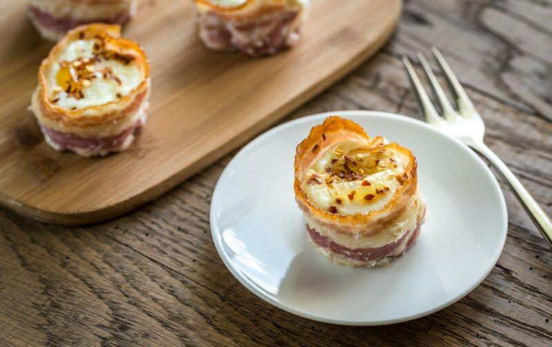 A small white plate with an egg cup made using bacon, next to a wooden board with additional egg cups