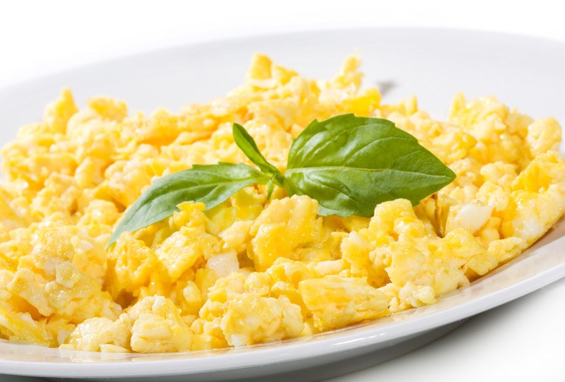 A white ceramic plate is filled with yellow scrambled eggs and a sprig of green leaves against a white background.