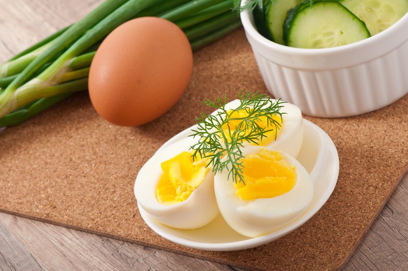 This photo shows three boiled egg halves on a small white dish with a green herb garnish, next to an egg, some green onions, and some cucumber slices in a white dish on a sheet of cork, which is on a wooden table.