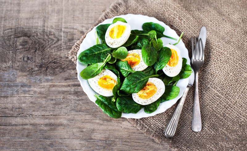 This photo shows boiled egg halves and spinach leaves on a white plate, lying next to a knife and fork on a beige burlap cloth on a wooden table.