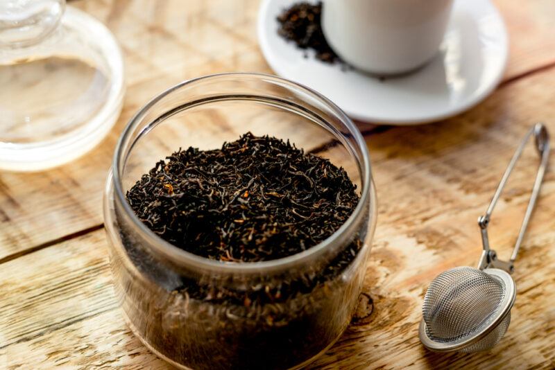 on a wooden surface is a small jar of loose English breakfast tea with its cover, a snap ball tea strainer, and a cup and saucer beside it