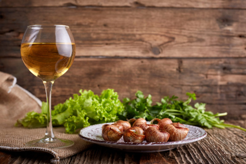 A white plate of escargot next to a glass of white wine and some greenery