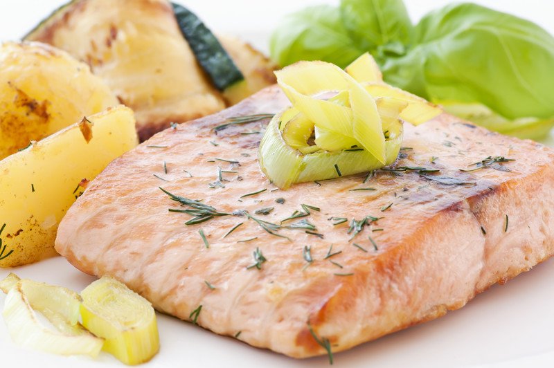 This photo shows a cooked salmon steak with onions, greens, and potato wedges on a white plate.