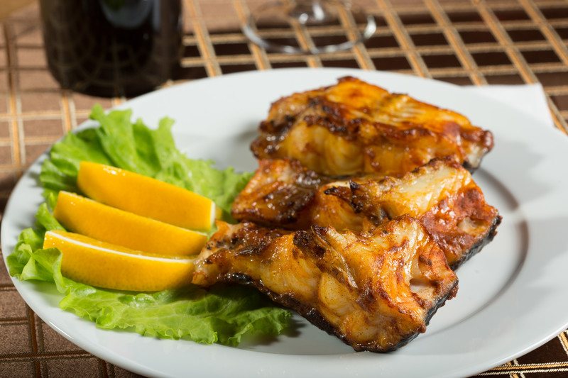 This photo shows a white plate with broiled fish, lemon wedges, and lettuce.