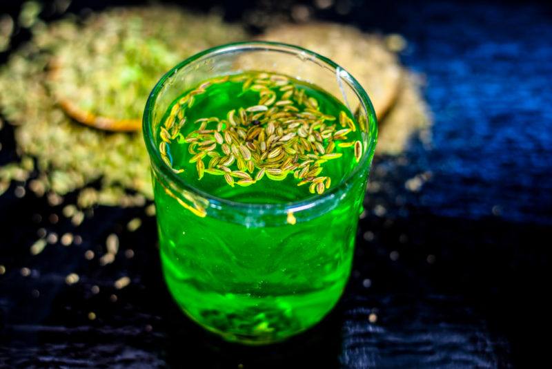 A glass of green water with fennel seeds on the top and in the background