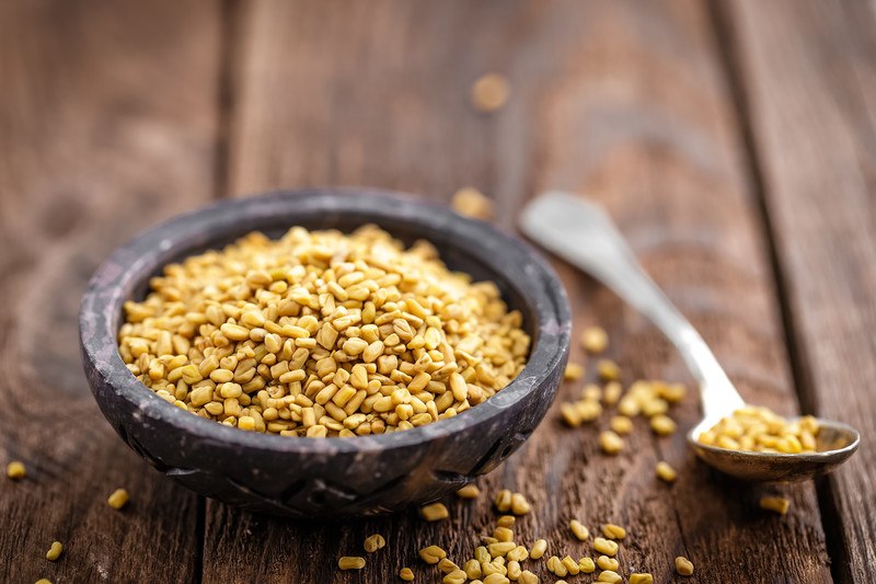 Several fenugreek seeds fill a dark bowl lying next to a spoon on a wooden table.