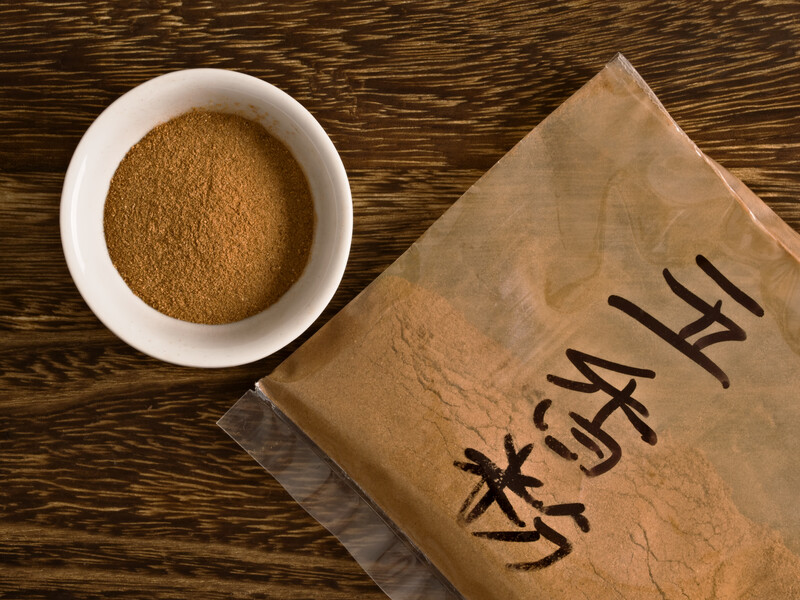 On top of a wooden surface, a small white bowl with five-spice powder beside a plastic envelope with Chinese writings, half-filled with five-spice powder. 