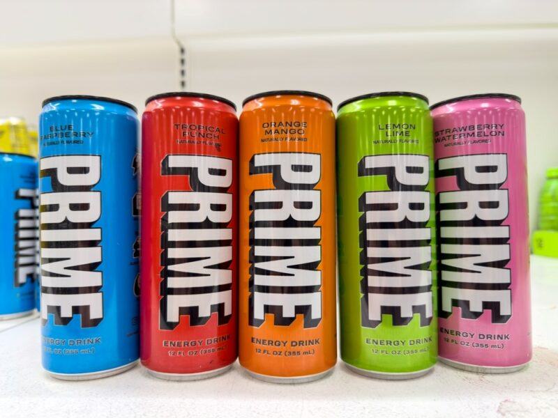 Cans of Prime energy drink, including Blue Raspberry, Tropical Punch, Orange Mango, Lemon Lime, and Strawberry Watermelon flavors.