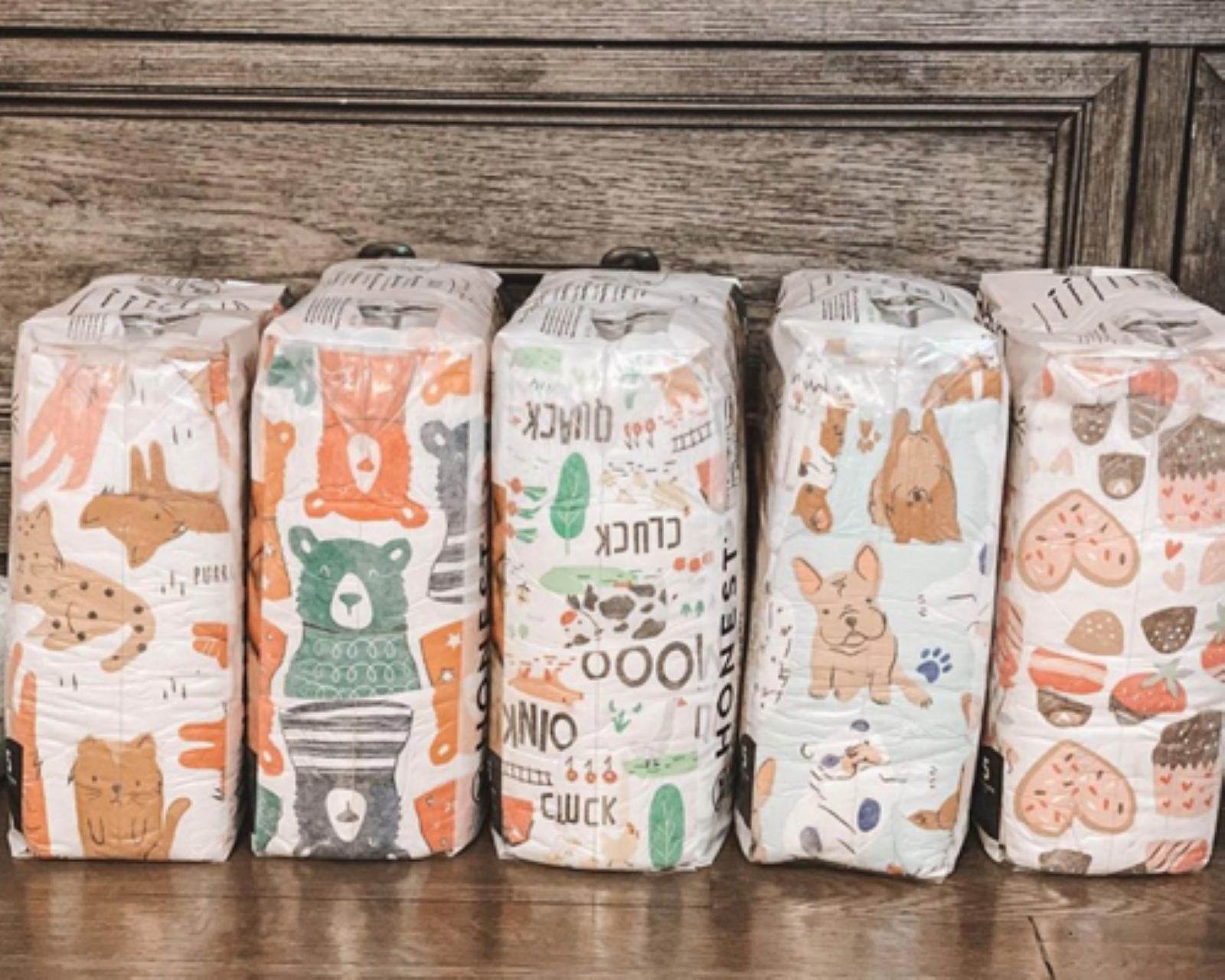 Five packages of diapers with cartoon decorations like  cats, bears, famr animals, dogs, hearts and desserts