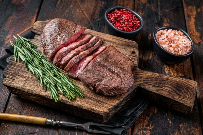 A wooden board on a table with a sliced piece of flat iron steak, rosemary sprigs, red peppercorns, and Himalyan salt