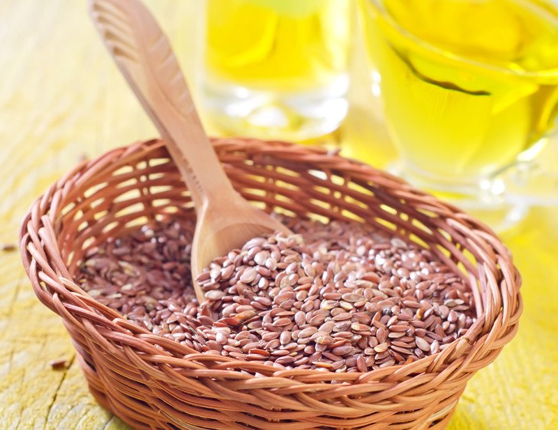 This photo shows a wicker basket filled with flax seeds and a wooden spoon in front of some glasses of yellow liquid on a wooden tabletop.