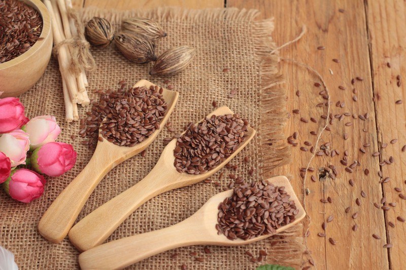 Three wooden spoons filled with flax seeds rest on a piece of burlap on a wooden table next to a bowl of flax seeds and five pink roses.