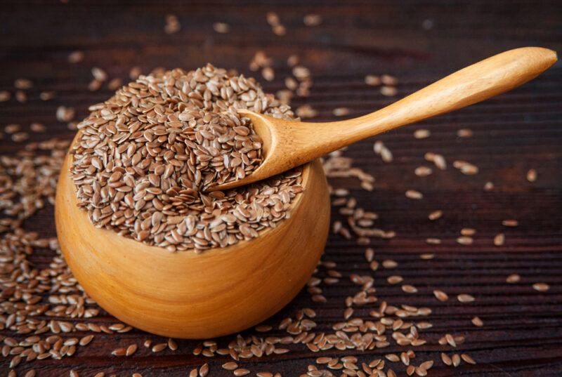 A wooden bowl ad wooden spoon are filled with flax seeds, next to loose flax seeds on a dark wooden surface.