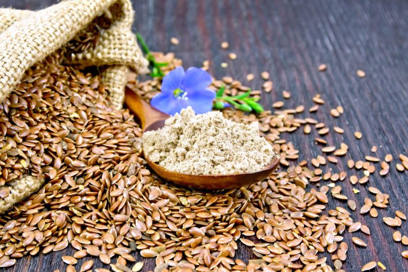 A wooden table with flax seeds spilling from a bag and a spoon of flaxseed meal