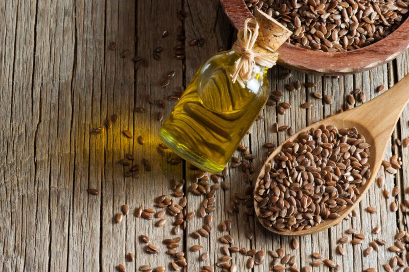 A large bowl and wooden spoon containing flax seeds, next to a bottle of flaxseed oil, with the seeds scattered across the table