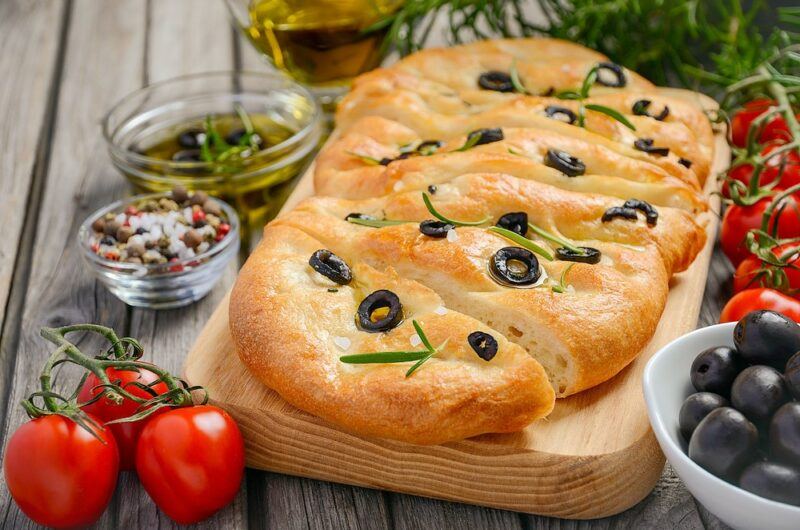 A wooden board with fresh rosemary and olive focaccia bread, next to oil, tomatoes, olives, and other ingredients