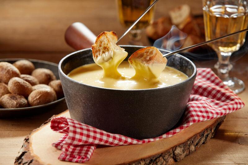 Two toasted baguettes on steel kabob sticks are being dipped into a cheese fondue in a dark pan that's resting on a red and white cloth, on a rustic piece of wood on a wooden tabletop.