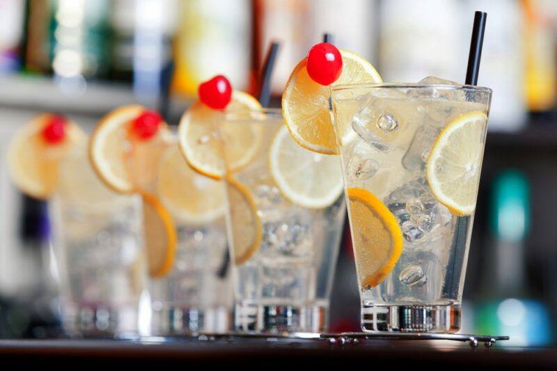 A line of Tom Collins cocktails on a bar, each garnished with lemon slices and a glace cherry