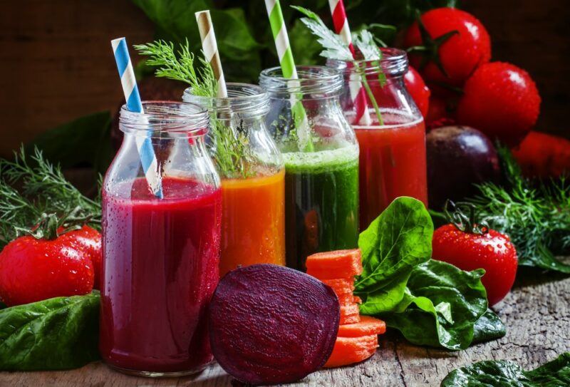 Four bottles of fruit juice, one pink, one orange, one green, one red, with various fresh vegetables on the table