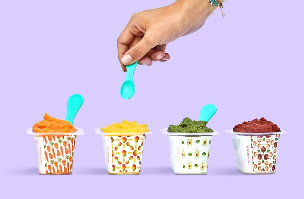 Four containers of baby food, from left to right carrot, mango, avocado, and beet  with a hand hovering over the mango baby food with a small blue spoon