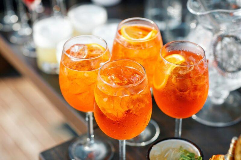 Four glasses containing an aperol spritz to be served at a party, with more glassware in the background