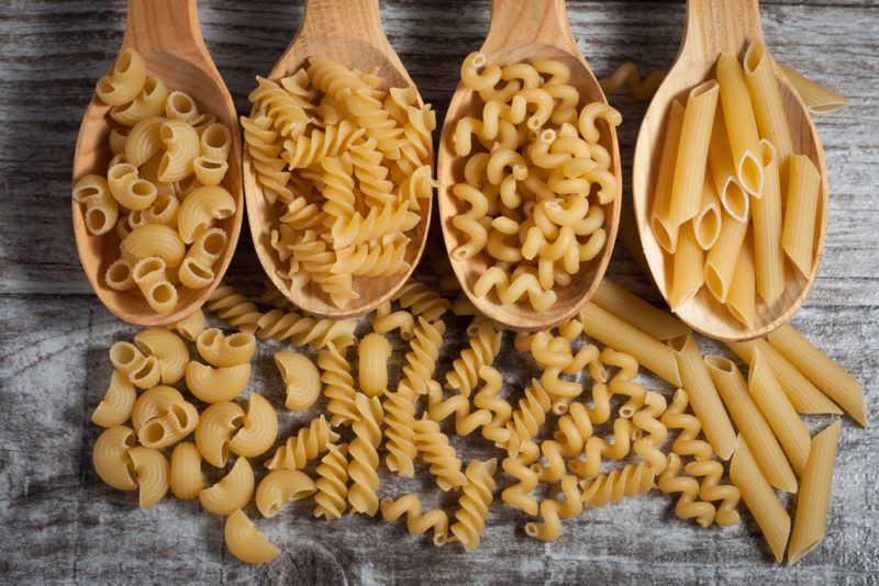 Four large wooden spoons with different types of pasta and the same types of pasta on a table