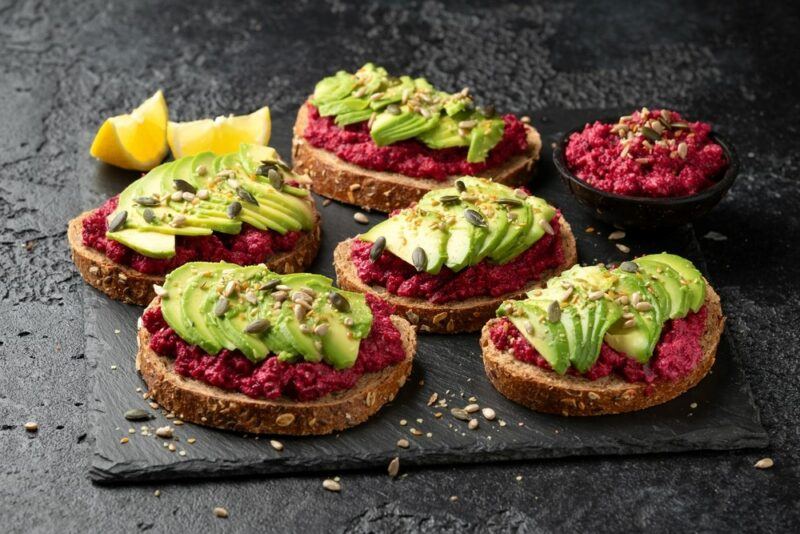 Five slices of sourdough, each topped with beetroot hummus, avocado, and seeds, with a bowl of beetroot hummus and some lemon wedges in the background