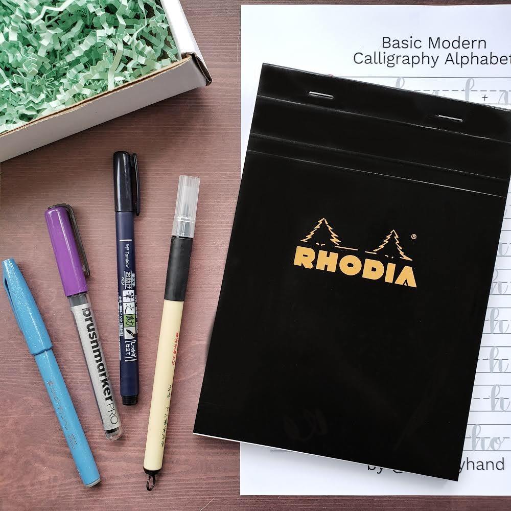 Four pens sitting to the left of a black covered notepad with gold wring of a silhouette of a two pines and RHODIA written below