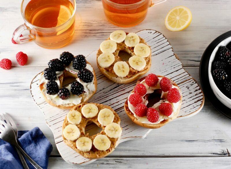 Four bagels lying flat on a board, each covered with spread and fresh fruit