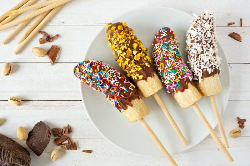 A white plate with four bananas, which are each dipped in chocolate and various toppings