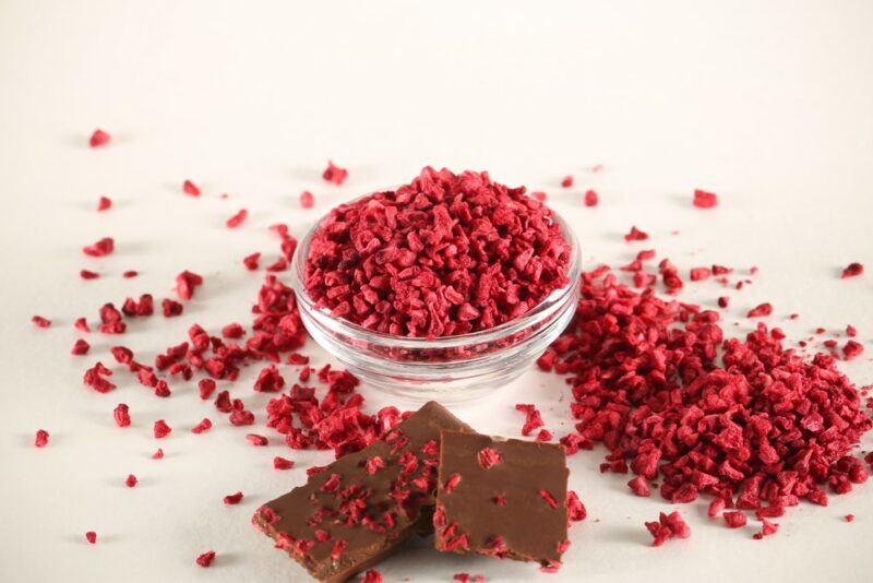 A small glass bowl of freeze dried raspberries, with more on the table, next to some chocolate