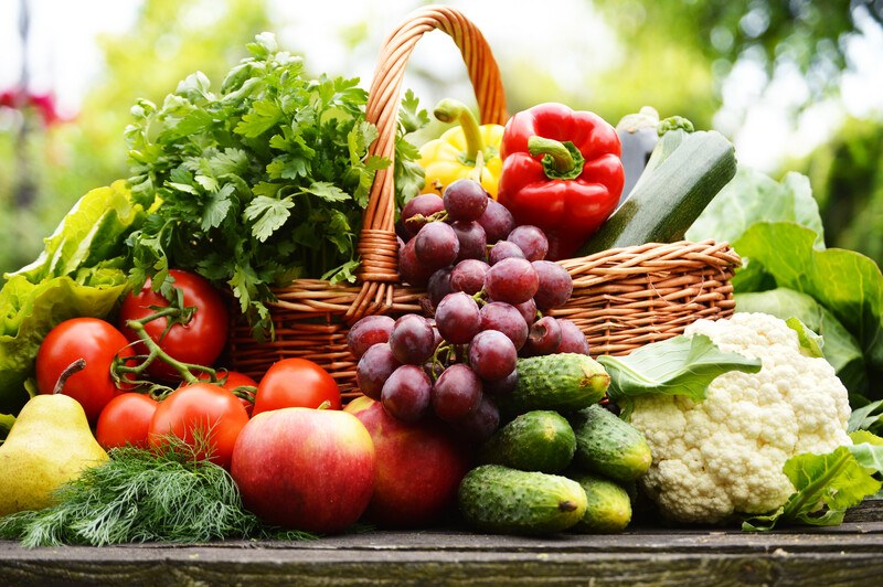 Basket full of vegetables fruits and herbs sitting on a table outside surrounded by more vegetables and fruit, including cucumbers, cauliflower, parsley, red peppers, tomatoes, and lettuce