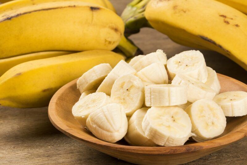 A wooden bowl with banana slices, in front of various yellow bananas