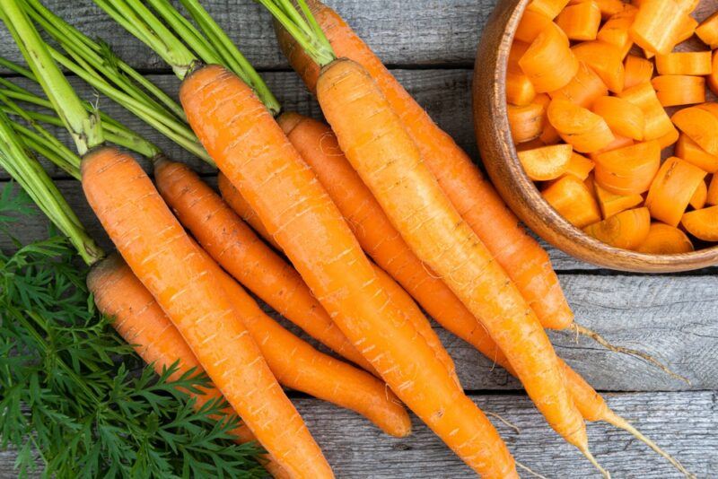 Fresh carrots lying flat on a wooden table, next to a bowl of chopped carrots