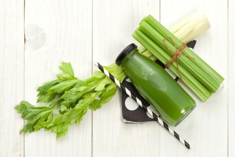 A white wooden table with a celery stalk, a bundle of smaller pieces of celery, a bottle of celery juice, and a straw