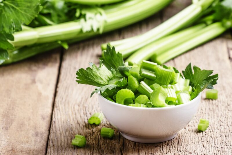 A wooden table with fresh celery stalks, next to a white bowl of chopped celery pieces