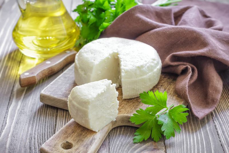 A wooden cutting board with a whole piece of fresh cheese that has a piece cut out of it. There is a cloth, herbs, and some oil in the background.